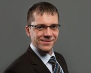 Ein Mann mit kurzen Haaren, Brille und dunklem Anzug, weißem Hemd und gestreifter Krawatte lächelt sanft vor einem grauen Hintergrund und verkörpert den professionellen Geist der Hochschule Coburg.