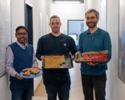 Drei lächelnde Personen stehen in einem Flur der Hochschule Coburg und halten selbst gebackene Backwaren in den Händen. Die Person links hat einen Teller mit Keksen, die Person in der Mitte hält ein Tablett mit Riegeln und die Person rechts hat ein Tablett mit Gebäck mit Erdbeeren.