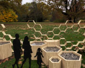 Die Silhouetten einer Familie, bestehend aus zwei Erwachsenen und einem Kind, stehen in einem Park in der Nähe der Hochschule Coburg neben einer Ausstellung sechseckiger, mit Kies gefüllter, an Waben erinnernder Pflanzkübel aus Holz, im Hintergrund sind verstreute Blätter und große Bäume zu sehen.