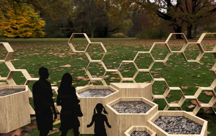 Die Silhouetten einer Familie, bestehend aus zwei Erwachsenen und einem Kind, stehen in einem Park in der Nähe der Hochschule Coburg neben einer Ausstellung sechseckiger, mit Kies gefüllter, an Waben erinnernder Pflanzkübel aus Holz, im Hintergrund sind verstreute Blätter und große Bäume zu sehen.
