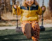 Ein Kind mit lockigem Haar und einer Beanie-Mütze sitzt auf einer Schaukel und hat den Fuß nach vorne gestreckt. Es trägt eine gelbe Jacke und orange gepunktete Hosen, was an den lebhaften Studentengeist der Hochschule Coburg erinnert. Im Hintergrund sind Bäume unter einem strahlend blauen Himmel zu sehen.