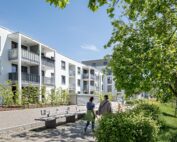 Unweit der Hochschule Coburg steht unter strahlend blauem Himmel ein moderner Wohnkomplex mit weißen Fassaden und Balkonen. Zwei Menschen schlendern einen von Bäumen und Bänken gesäumten Weg entlang, der eine ruhige und einladende Atmosphäre schafft.