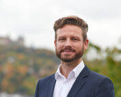 Ein Mann mit Vollbart und Schnurrbart steht im Freien auf dem Campus der Hochschule Coburg. Er trägt ein weißes Hemd und einen dunklen Blazer. Im Hintergrund ist eine verschwommene Landschaft mit Bäumen und einem entfernten Gebäude zu sehen. Er lächelt leicht und blickt in die Kamera.