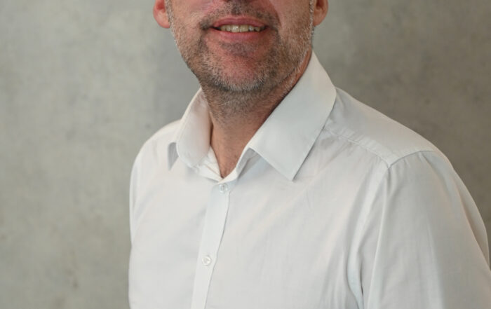 Ein Mann mit kurzen dunklen Haaren und Bart, der ein weißes Hemd mit Knöpfen trägt, steht vor einem schlichten grauen Hintergrund. Mit einem sanften Lächeln verkörpert er den professionellen Geist der Hochschule Coburg, während er direkt in die Kamera blickt.