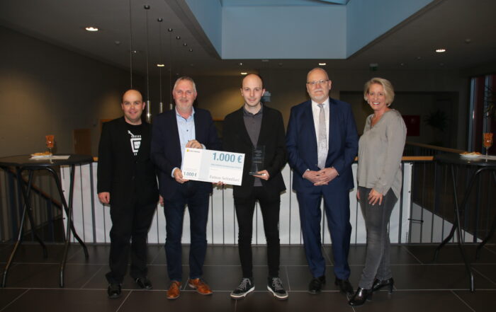 Eine Gruppe von fünf Personen steht drinnen, in der Mitte hält eine Person ein Zertifikat und einen großen Scheck über 1.000 Euro. Als Vertreter der Hochschule Coburg sind alle in Business- oder eleganter Freizeitkleidung gekleidet und alle lächeln strahlend.