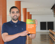 Eine Person im blauen Poloshirt steht in einem Flur der Hochschule Coburg und hält ein Buch mit dem Titel "Kosteneffizienz und nachhaltige Automobilie" in der Hand. Im Hintergrund sind orangefarbene Säulen und eine Treppe zu sehen, die eine akademische Atmosphäre schaffen.