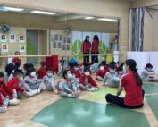 Eine Gruppe kleiner Kinder in passenden rot-grauen Uniformen sitzt auf dem polierten Holz- und grün gefliesten Boden der Turnhalle der Hochschule Coburg. Sie stehen einem Lehrer gegenüber, tragen alle Masken und sitzen neben einer Spiegelwand, die ihre aufmerksamen Ausdrücke widerspiegelt.