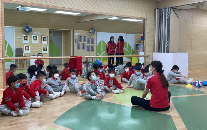 Eine Gruppe kleiner Kinder in passenden rot-grauen Uniformen sitzt auf dem polierten Holz- und grün gefliesten Boden der Turnhalle der Hochschule Coburg. Sie stehen einem Lehrer gegenüber, tragen alle Masken und sitzen neben einer Spiegelwand, die ihre aufmerksamen Ausdrücke widerspiegelt.