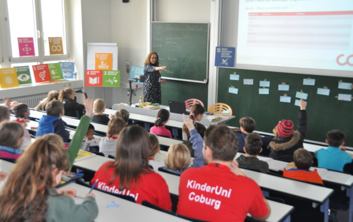 Eine Frau von der Hochschule Coburg hält einen Vortrag vor Kindern in einem Klassenraum. Die Schüler sitzen an ihren Tischen, einige heben die Hand. An den Wänden hängen Plakate mit Zielen für nachhaltige Entwicklung, und auf der Projektionsfläche wird eine Folie mit Text und einer Grafik gezeigt.