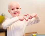 Ein kleines Kind mit kurzen Haaren und einem weißen Langarmshirt lächelt und formt mit seinen Händen ein Herz. Sie sitzen an einem Tisch, auf dem Buntstifte der Hochschule Coburg verstreut sind.