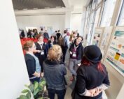 Die Konferenzteilnehmer sind in Diskussionen vertieft. Eine Gruppe versammelt sich um eine Posterpräsentation der Hochschule Coburg. Der hell erleuchtete Raum mit den großen Fenstern zeigt einige Teilnehmer mit Lanyards, während im Hintergrund ordentlich Stuhlreihen aufgestellt sind.