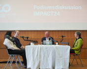 Vier Personen sitzen an einem Tisch und diskutieren an einer Podiumsdiskussion bei der Veranstaltung „Podiumsdiskussion IMPACT24“, die von der Hochschule Coburg veranstaltet wird. Mikrofone und Wasserflaschen schmücken den Tisch, während im Hintergrund ein großes Schild mit der Aufschrift „Hochschule Coburg“ zu sehen ist.