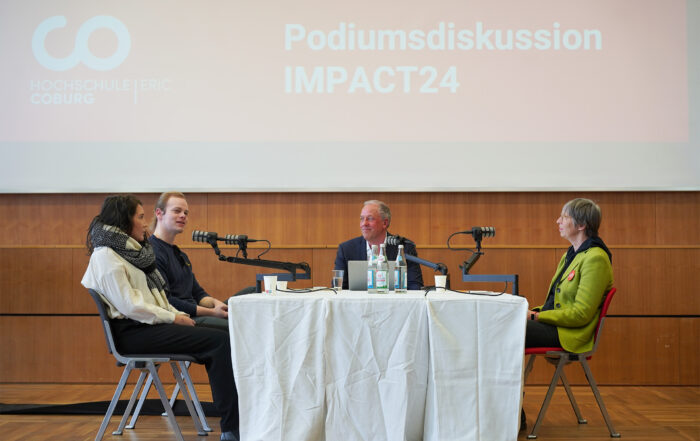 Vier Personen sitzen an einem Tisch und diskutieren an einer Podiumsdiskussion bei der Veranstaltung „Podiumsdiskussion IMPACT24“, die von der Hochschule Coburg veranstaltet wird. Mikrofone und Wasserflaschen schmücken den Tisch, während im Hintergrund ein großes Schild mit der Aufschrift „Hochschule Coburg“ zu sehen ist.