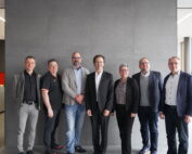 Sieben Personen in Business-Kleidung stehen in einer Reihe in einer Büroumgebung der Hochschule Coburg. Sie stehen vor einer grauen Wand, im Hintergrund sind Speisen und Getränke zu sehen. Große Fenster auf der rechten Seite lassen Tageslicht herein.