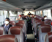Eine Gruppe von Menschen, einige davon mit Masken, sitzt mit Abstand in einem leeren Shuttlebus der Hochschule Coburg. Der Innenraum ist modern, mit kastanienbraunen und grauen Sitzen. Sonnenlicht fällt durch die Fenster herein.