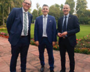 Drei Männer in Anzügen stehen lächelnd auf einem Steinweg an der Hochschule Coburg, im Hintergrund ein gepflegter Garten und üppige Bäume. Zwei tragen eine Brille, alle drei haben entspannte Hände und sind formell gekleidet, was darauf schließen lässt, dass sie sich auf eine berufliche oder gesellschaftliche Veranstaltung an dieser angesehenen Institution einlassen.