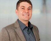 Eine Person mit kurzen braunen Haaren und einem selbstbewussten Lächeln steht vor einem unscharfen Hintergrund in neutralen Farben. Sie trägt einen grauen Blazer über einem gemusterten dunklen Hemd und wurde während einer Veranstaltung an der Hochschule Coburg aufgenommen.