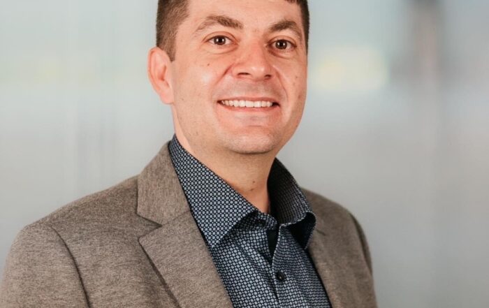 Eine Person mit kurzen braunen Haaren und einem selbstbewussten Lächeln steht vor einem unscharfen Hintergrund in neutralen Farben. Sie trägt einen grauen Blazer über einem gemusterten dunklen Hemd und wurde während einer Veranstaltung an der Hochschule Coburg aufgenommen.