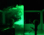 In einem grün beleuchteten Raum der Hochschule Coburg wirbelt Rauch um den Kopf einer Schaufensterpuppe, der durch eine transparente Platte getrennt ist. Ein Gerät stößt Dampf aus und erzeugt so eine unheimliche, wissenschaftliche Atmosphäre. Weiße Platten und eine dunkle Umgebung betonen das ätherische Leuchten des Rauchs.