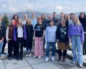 Eine Gruppe von Menschen steht im Freien auf einer gepflasterten Fläche der Hochschule Coburg, im Hintergrund eine Hügellandschaft und ein Gebäude. In legerer Kleidung lächeln sie an diesem sonnigen Tag in die Kamera.