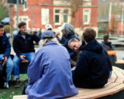 Eine Gruppe von Menschen sitzt auf geschwungenen Holzbänken im Freien vor der Hochschule Coburg und unterhält sich. Warm gekleidet unterhalten sie sich in einer lockeren und geselligen Umgebung mit Gebäuden, Bäumen und einem geparkten Auto im Hintergrund.