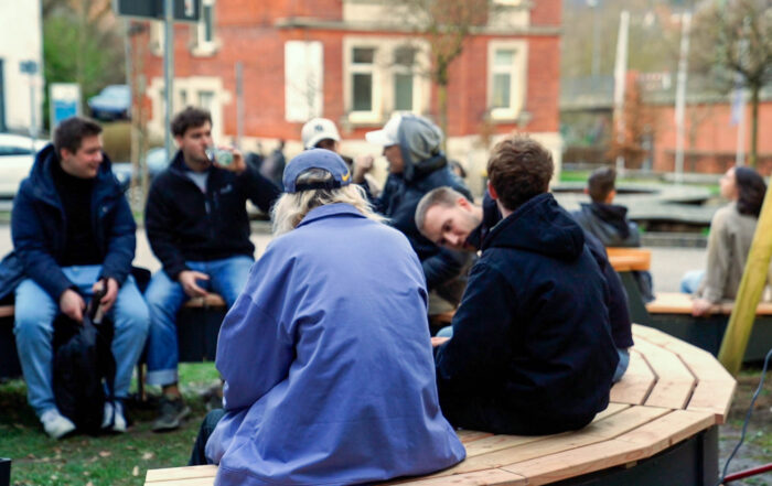 Eine Gruppe von Menschen sitzt auf geschwungenen Holzbänken im Freien vor der Hochschule Coburg und unterhält sich. Warm gekleidet unterhalten sie sich in einer lockeren und geselligen Umgebung mit Gebäuden, Bäumen und einem geparkten Auto im Hintergrund.