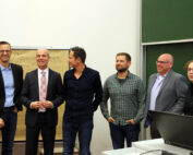 Eine Gruppe von sechs professionell gekleideten Personen steht in einem Unterrichtsraum der Hochschule Coburg. Einige lächeln. Auf einem Tisch steht ein Laptop, im Hintergrund ein Flipchart und eine große Tafel.