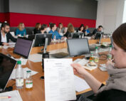 Eine Gruppe von Menschen sitzt an einem langen Konferenztisch in der Hochschule Coburg und ist in eine Besprechung vertieft. Einige konzentrieren sich auf Laptops und Dokumente. Eine Frau im Vordergrund hält eine ausgedruckte Tagesordnung in der Hand. Auf dem Tisch stehen mehrere Computermonitore und Getränkeflaschen.