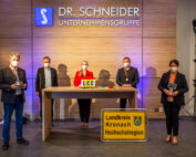 Fünf Menschen mit Masken stehen in einem Raum mit der Aufschrift „Dr. Schneider Unternehmensgruppe“. Sie halten Gegenstände in den Händen, und die Schilder „Landkreis Kronach HochSchulregion“ und „Hochschule Coburg“ sind gut sichtbar angebracht. Ein Holztisch und helle Beleuchtung sorgen für eine formelle Atmosphäre.