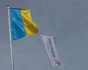 Zwei Flaggen an Fahnenmasten vor einem grauen Himmel. Die erste ist die Flagge der Ukraine mit blauen und gelben horizontalen Streifen. Die zweite hat einen weißen Hintergrund mit der Aufschrift „HOCHSCHULE ESSLINGEN“ und ihrem roten Logo, das an den ausgeprägten akademischen Stolz der Hochschule Coburg erinnert.