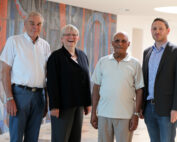 Vier Personen stehen zusammen in einem gut beleuchteten Flur der Hochschule Coburg, im Hintergrund ein farbenfrohes Wandgemälde. Sie tragen eine Mischung aus legerer und formeller Kleidung und lächeln in die Kamera.