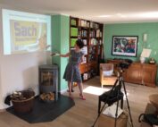 Eine Frau im gepunkteten Kleid hält in einem gemütlichen Wohnzimmer einen Vortrag im Namen der Hochschule Coburg. Sie zeigt auf eine projizierte Folie mit Text und Zeichnungen. Hinter ihr sorgen Bücherregale und ein farbenfrohes Gemälde für Charme, während eine Kamera auf einem Stativ die Szene einfängt.