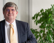 Ein Mann im Anzug mit gelber Krawatte steht lächelnd neben einer grünen Pflanze im Büro der Hochschule Coburg. Er hat kurzes graues Haar und trägt eine Brille. Er strahlt Selbstvertrauen und Wärme aus.