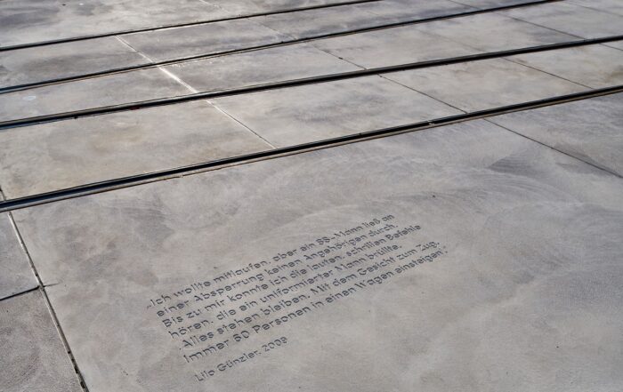 In den Beton neben den Straßenbahnschienen ist ein deutsches Zitat eingraviert, das auf das Warten auf ein Signal zum sicheren Überqueren der Straße hinweist. Es wird Lilo Gunter zugeschrieben (2008). Der Bereich nahe der Hochschule Coburg ist in Sonnenlicht getaucht, was die Sichtbarkeit dieser nachdenklichen Erinnerung erhöht.