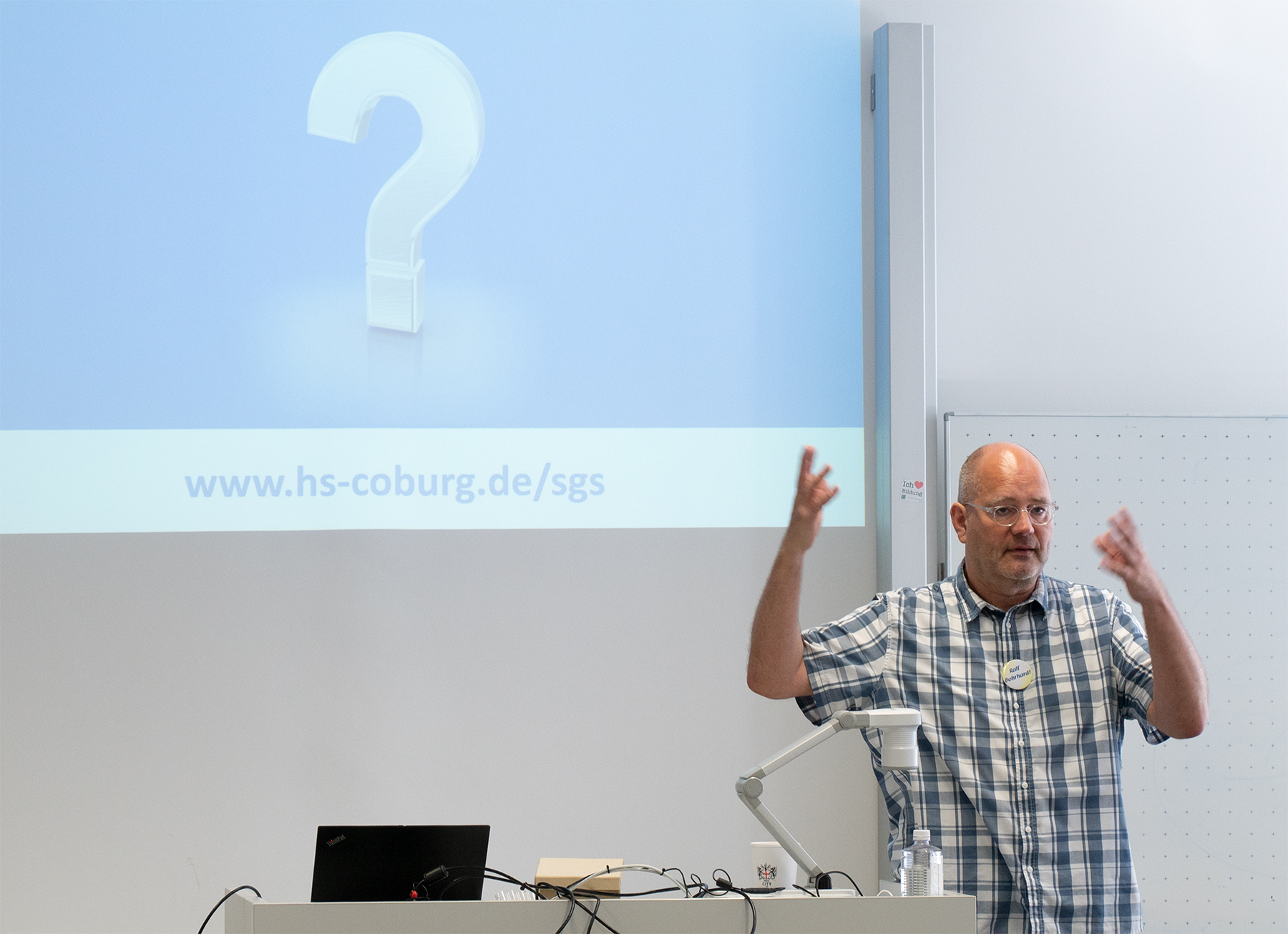 Ein Mann im karierten Hemd gestikuliert lebhaft während seines Vortrags an der Hochschule Coburg. Hinter ihm zeigt ein Bildschirm ein großes Fragezeichen und die URL: www.hs-coburg.de/sgs.