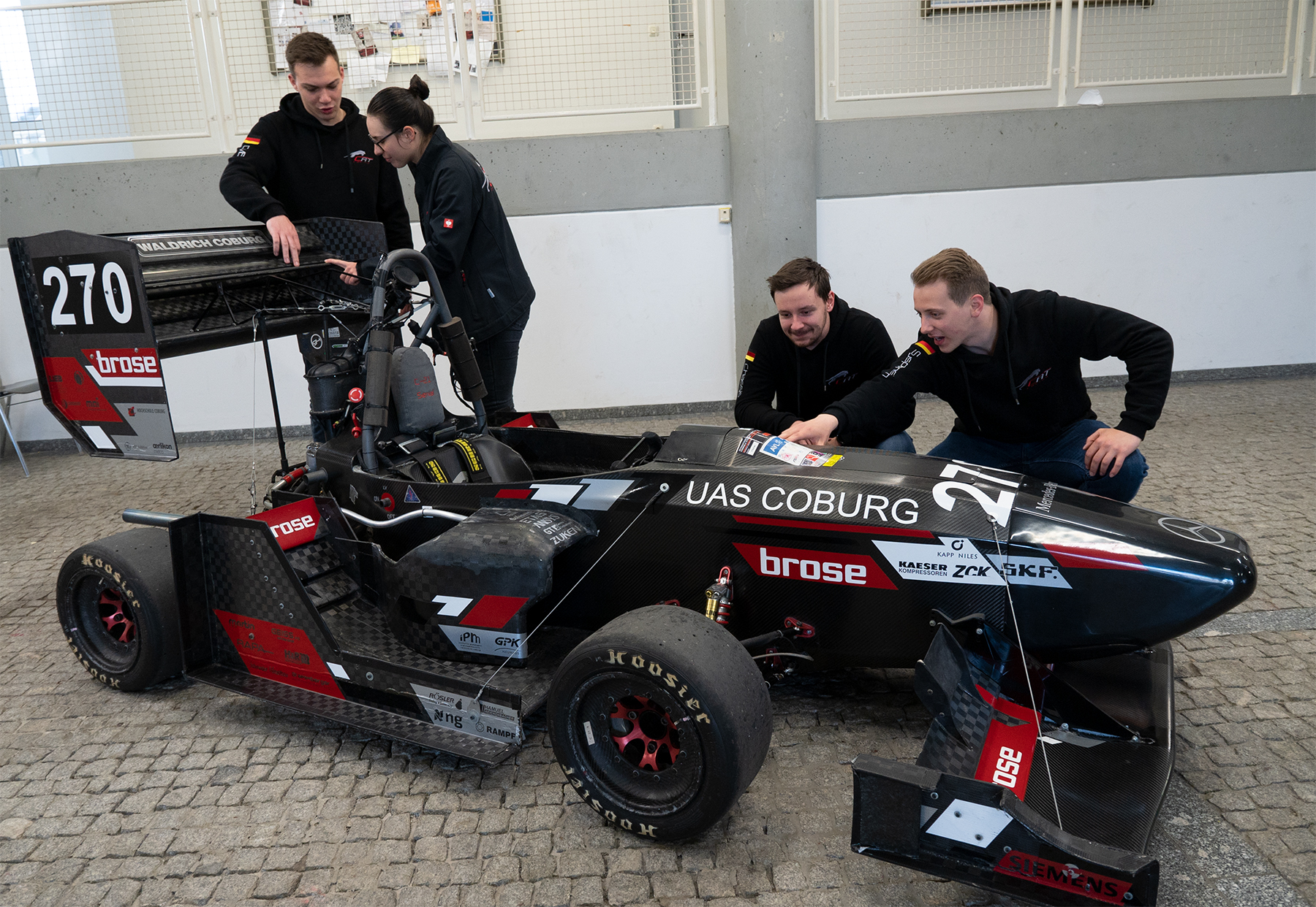 Vier Personen untersuchen einen schwarz-roten Formula Student-Rennwagen mit der Aufschrift „UAS Coburg“, der stolz die Hochschule Coburg repräsentiert. Das Auto, geschmückt mit verschiedenen Sponsorenlogos und der Nummer 270, steht in einer Werkstatt oder Garage mit gefliestem Boden, während sie sein elegantes Design begutachten.