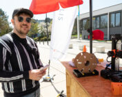 Eine Person mit Sonnenbrille und Kappe steht im Freien neben einem Tisch mit einem 3D-Drucker und einer Spule Filament an der Hochschule Coburg. Die sonnige Umgebung, eingerahmt von Bäumen und einem modernen Gebäude, ist perfekt, während sie ihr Smartphone hält.