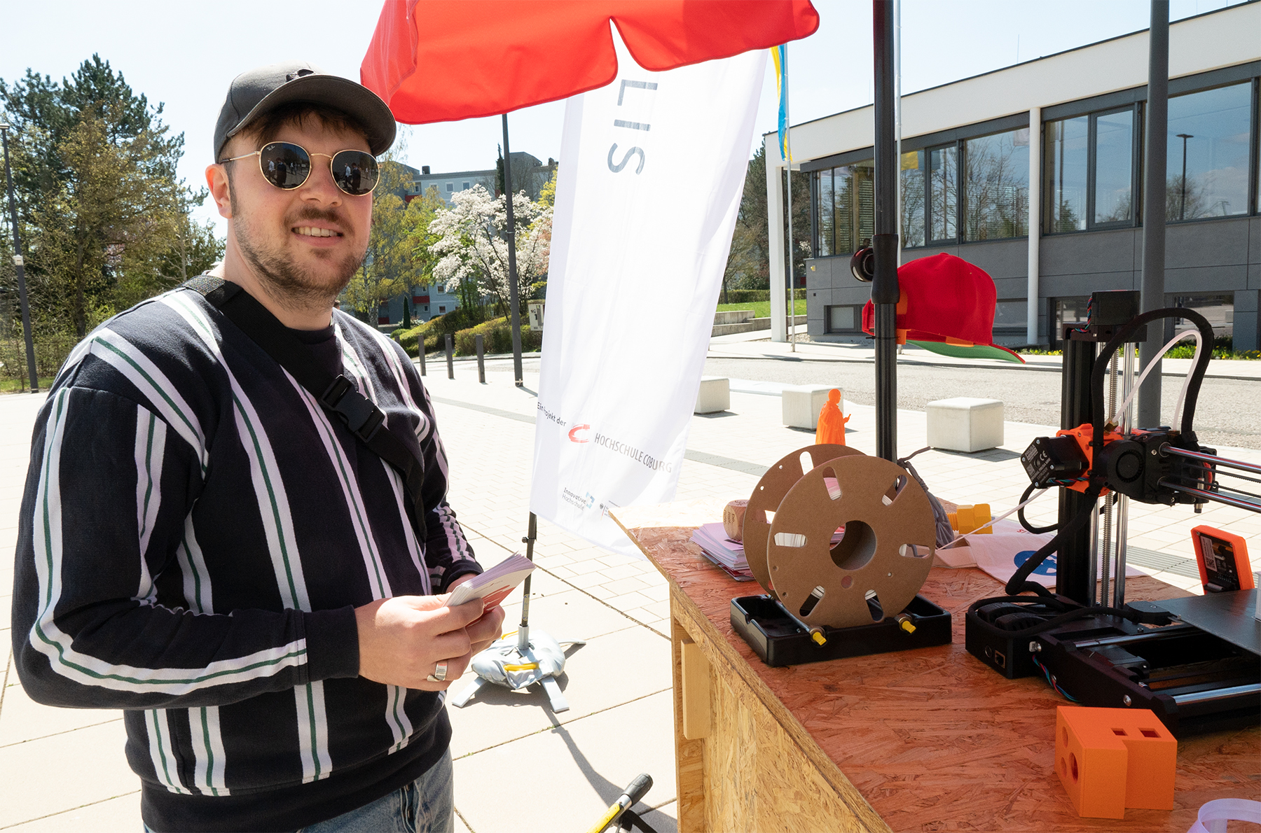 Eine Person mit Sonnenbrille und Kappe steht im Freien neben einem Tisch mit einem 3D-Drucker und einer Spule Filament an der Hochschule Coburg. Die sonnige Umgebung, eingerahmt von Bäumen und einem modernen Gebäude, ist perfekt, während sie ihr Smartphone hält.