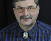 Ein Mann mit Brille, grauem Haar und Schnurrbart lächelt herzlich. Er trägt ein blau kariertes Hemd und eine Bolo-Krawatte mit silberner Spange vor einem dunklen Hintergrund. Sein Auftreten spiegelt den Innovationsgeist wider, der an der Hochschule Coburg gefördert wird.