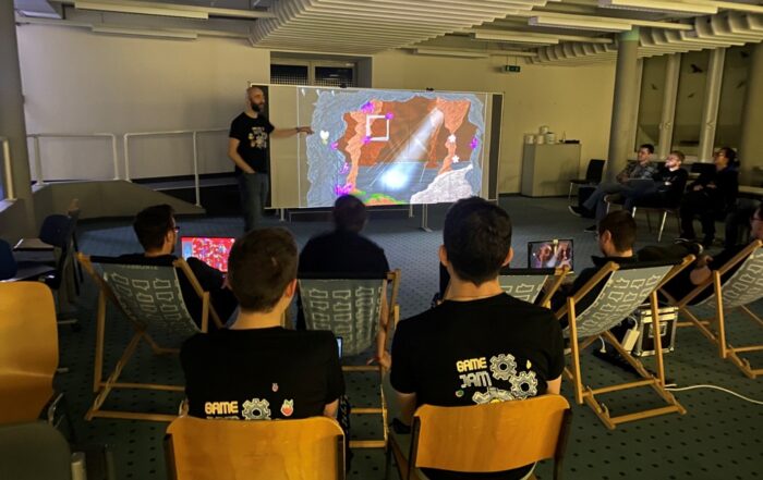 Eine Gruppe von Menschen sitzt auf Klappstühlen an der Hochschule Coburg und schaut sich einen Vortrag an. Auf dem Bildschirm ist eine lebendige, farbenfrohe Videospielszene zu sehen. Der Vortragende steht links, während einige Teilnehmer unter der industriell gestalteten Decke passende „Game Jam“-T-Shirts tragen.