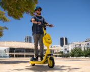 Bei klarem Himmel rast eine Person mit Sonnenbrille und Helm auf einem leuchtend gelben Elektroroller über einen Stadtplatz. Die Kulisse aus modernen Gebäuden und Bäumen lässt den lebendigen Geist der Hochschule Coburg erahnen.