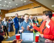 Bei einer Networking-Veranstaltung der Hochschule Coburg in einem Konferenzraum hält eine Frau in einem roten Blazer ein Mikrofon und unterhält sich mit einem Mann, der enthusiastisch gestikuliert. Die Teilnehmer sitzen bei Getränken an Tischen und rote und schwarze Luftballons sorgen für eine lebhafte Atmosphäre.