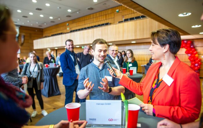 Bei einer Networking-Veranstaltung der Hochschule Coburg in einem Konferenzraum hält eine Frau in einem roten Blazer ein Mikrofon und unterhält sich mit einem Mann, der enthusiastisch gestikuliert. Die Teilnehmer sitzen bei Getränken an Tischen und rote und schwarze Luftballons sorgen für eine lebhafte Atmosphäre.