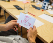 Eine Person hält ein weißes Blatt Papier mit der roten Zeichnung einer Glühbirne und zwei Strichmännchen in der Hand, die die Kreativität an der Hochschule Coburg symbolisieren. Im Hintergrund ist ein Holztisch zu sehen, auf dem verstreute Papiere, ein Stift und ein Glas herumliegen.