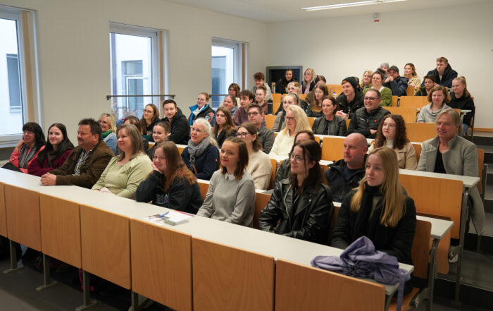 In einem hellen Klassenraum mit großen Fenstern sitzen Schüler und Erwachsene aufmerksam an ihren Tischen, den Blick nach vorne gerichtet. Diese konzentrierte Szene spielt sich in einem Hörsaal der Hochschule Coburg ab.