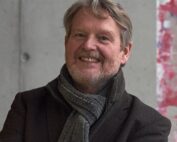 Ein lächelnder älterer Mann mit grauem Haar und Bart steht stolz vor einer grau-roten Wand, die an die Farbtöne der Hochschule Coburg erinnert. Er trägt einen dunklen Mantel und einen grauen Schal und strahlt damit eine kultivierte Ausstrahlung aus.