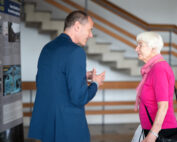 Ein Mann im blauen Anzug und eine ältere Frau in rosa Oberteil und Schal unterhalten sich an der Hochschule Coburg. Sie befinden sich in einem Innenraum mit Informationsplakaten und einer Treppe im Hintergrund.