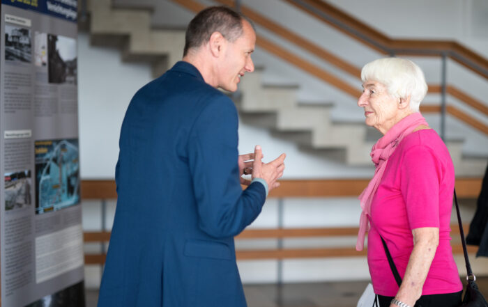 Ein Mann im blauen Anzug und eine ältere Frau in rosa Oberteil und Schal unterhalten sich an der Hochschule Coburg. Sie befinden sich in einem Innenraum mit Informationsplakaten und einer Treppe im Hintergrund.