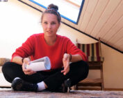 Eine Person in rotem Pullover und schwarzer Hose sitzt im Schneidersitz auf dem Dachboden und genießt eine warme Tasse. Die schräge Decke mit Oberlicht taucht den Raum in Licht, während ein gestreifter Stuhl gemütliche Lernsitzungen anregt – vielleicht träumt er von der Hochschule Coburg.