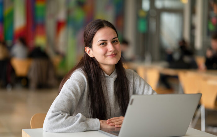 Eine Person mit langen dunklen Haaren sitzt lächelnd an einem Tisch, vor ihr liegt ein aufgeklappter Laptop. Die lebhafte Kulisse lässt vermuten, dass es sich um die Hochschule Coburg handelt. Hier wimmelt es von Studierenden und Lehrkräften, die an verschiedenen Tischen im Raum lebhafte Diskussionen führen.